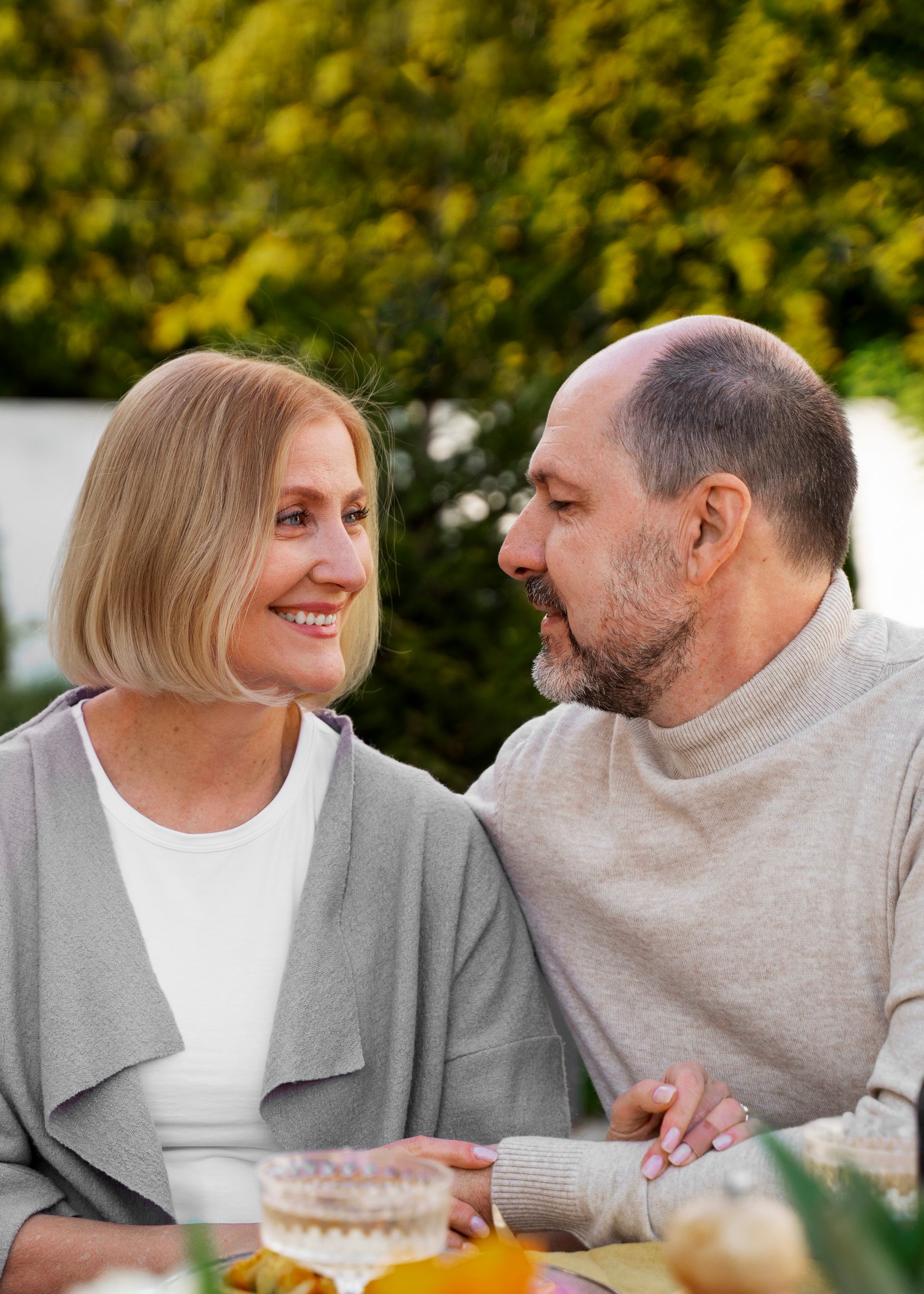 Smiling couple at an outdoor table, woman with blonde hair, man with a beard, foliage in background.