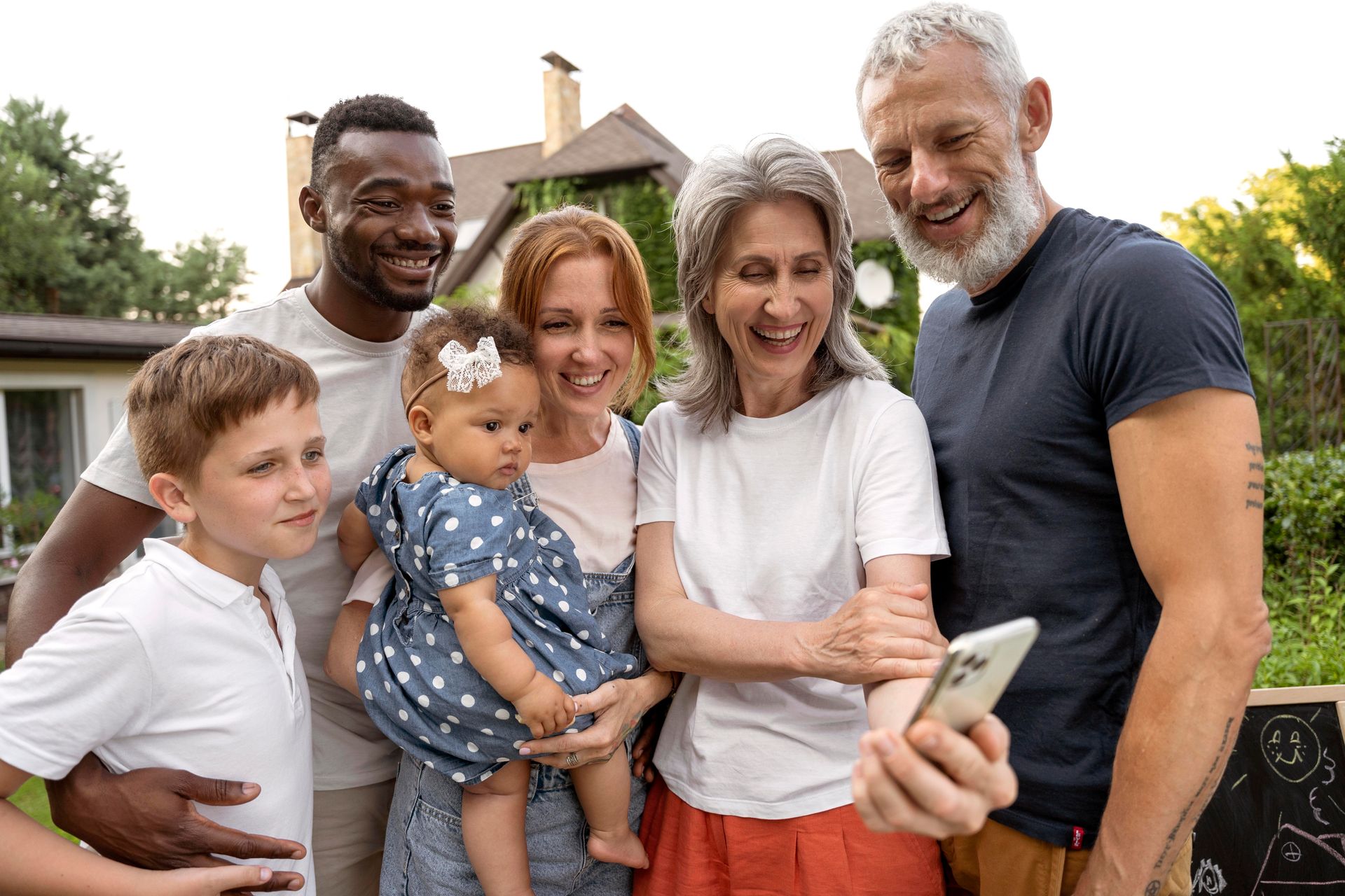 Family looking at a phone, includes a baby, in front of a house, smiling.