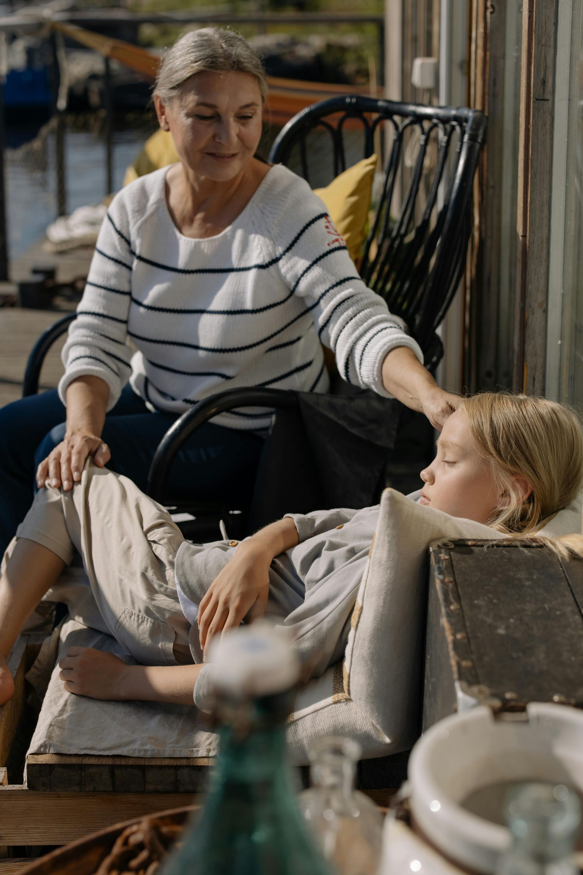 Woman comforts a sleeping child. Seated outdoors, she gently touches the child's head. Soft sunlight.