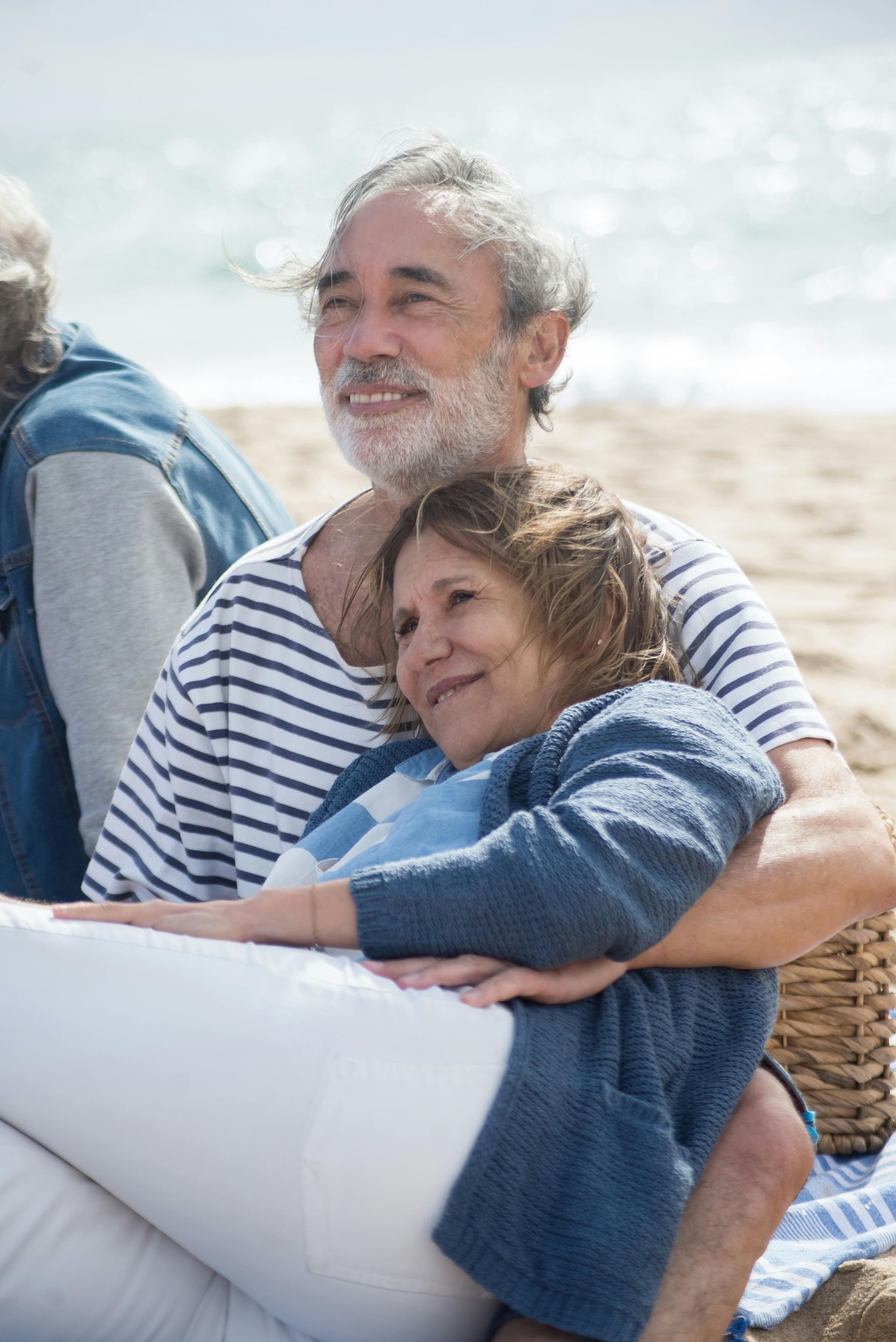 Elderly couple on a beach, man in stripes embraces woman, looking toward the sea.