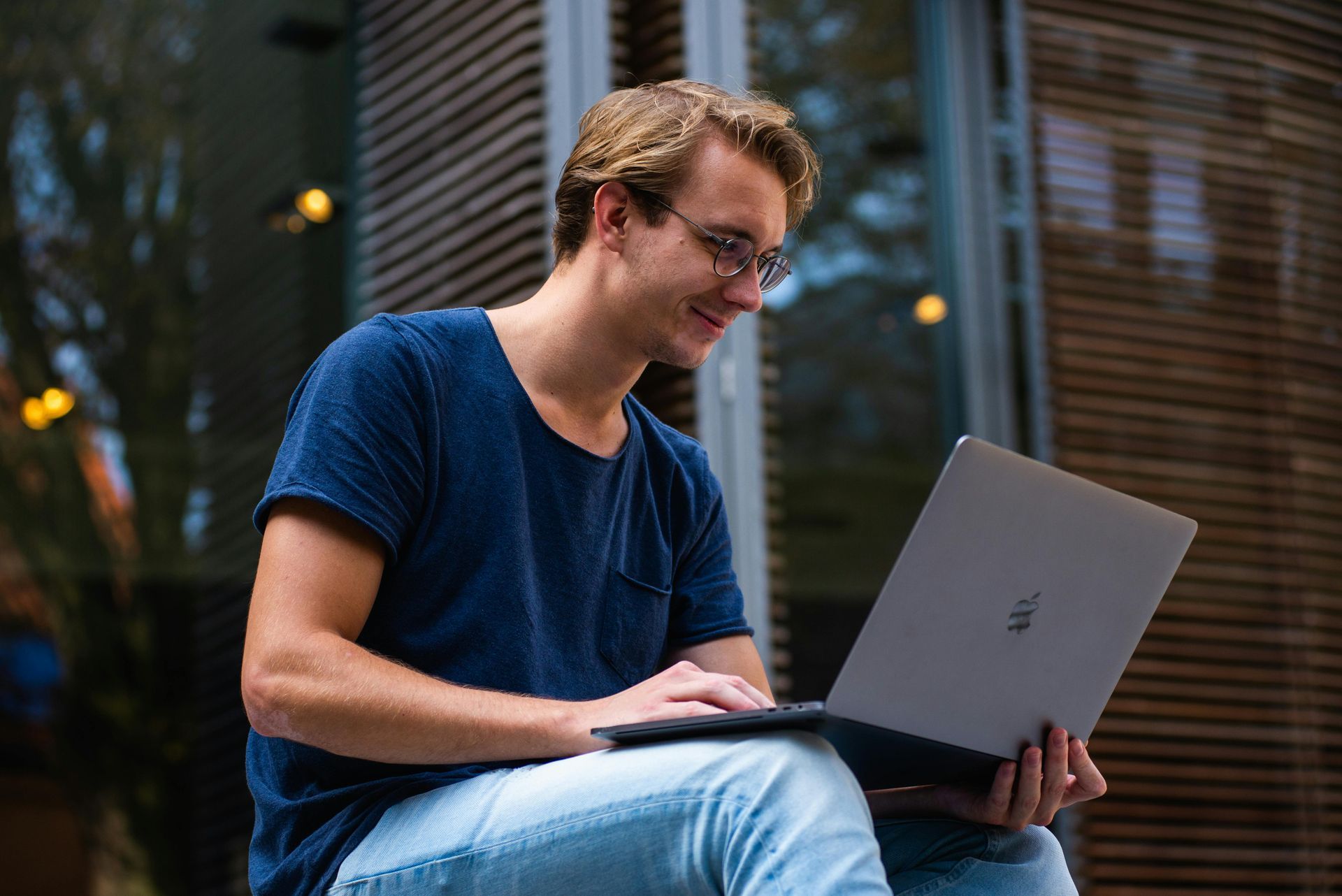 Man with blonde hair and glasses, smiling while using laptop outdoors.