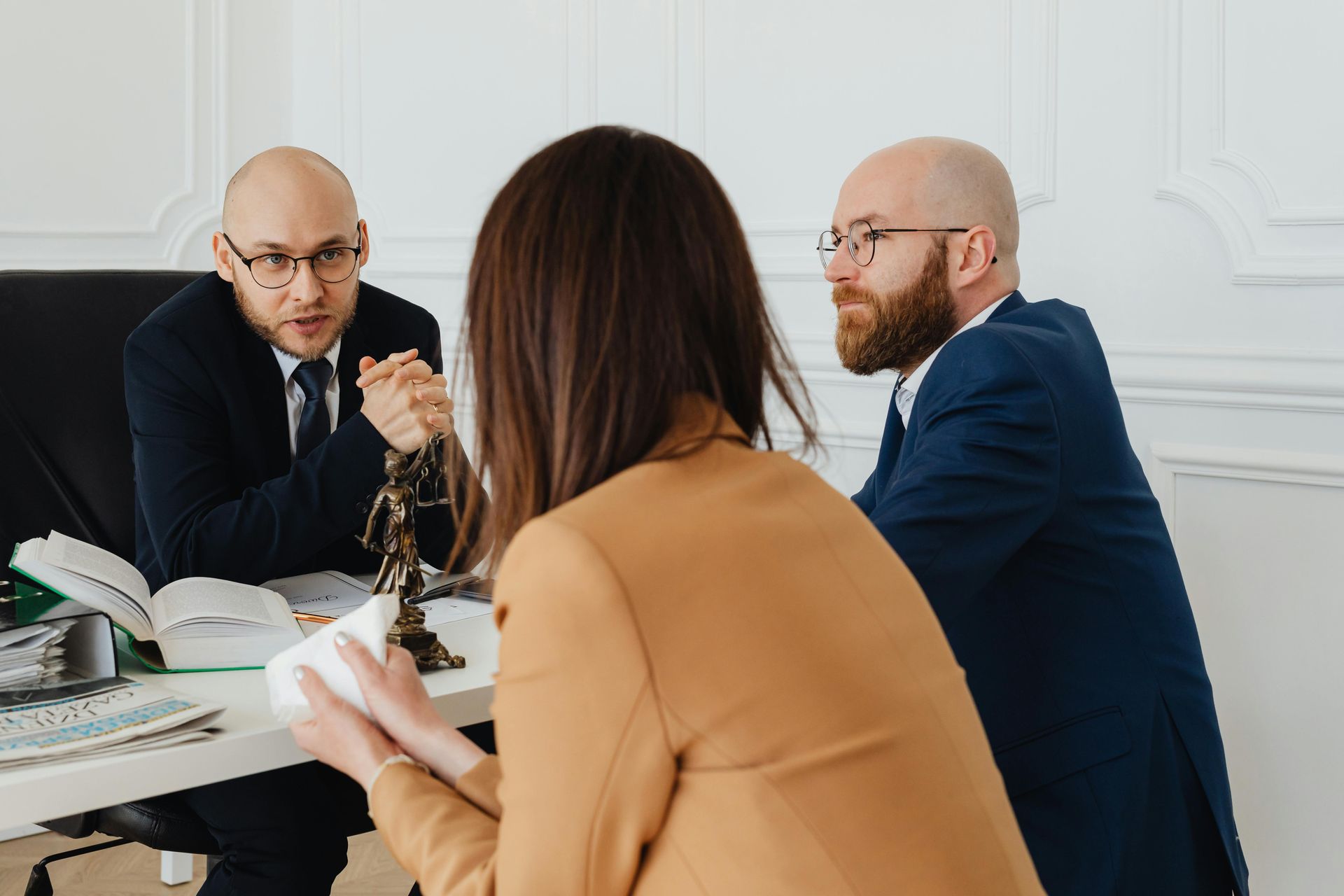 Three people in suits at a table: two men, one woman. Discussing, holding papers. Statuette, books, desk.