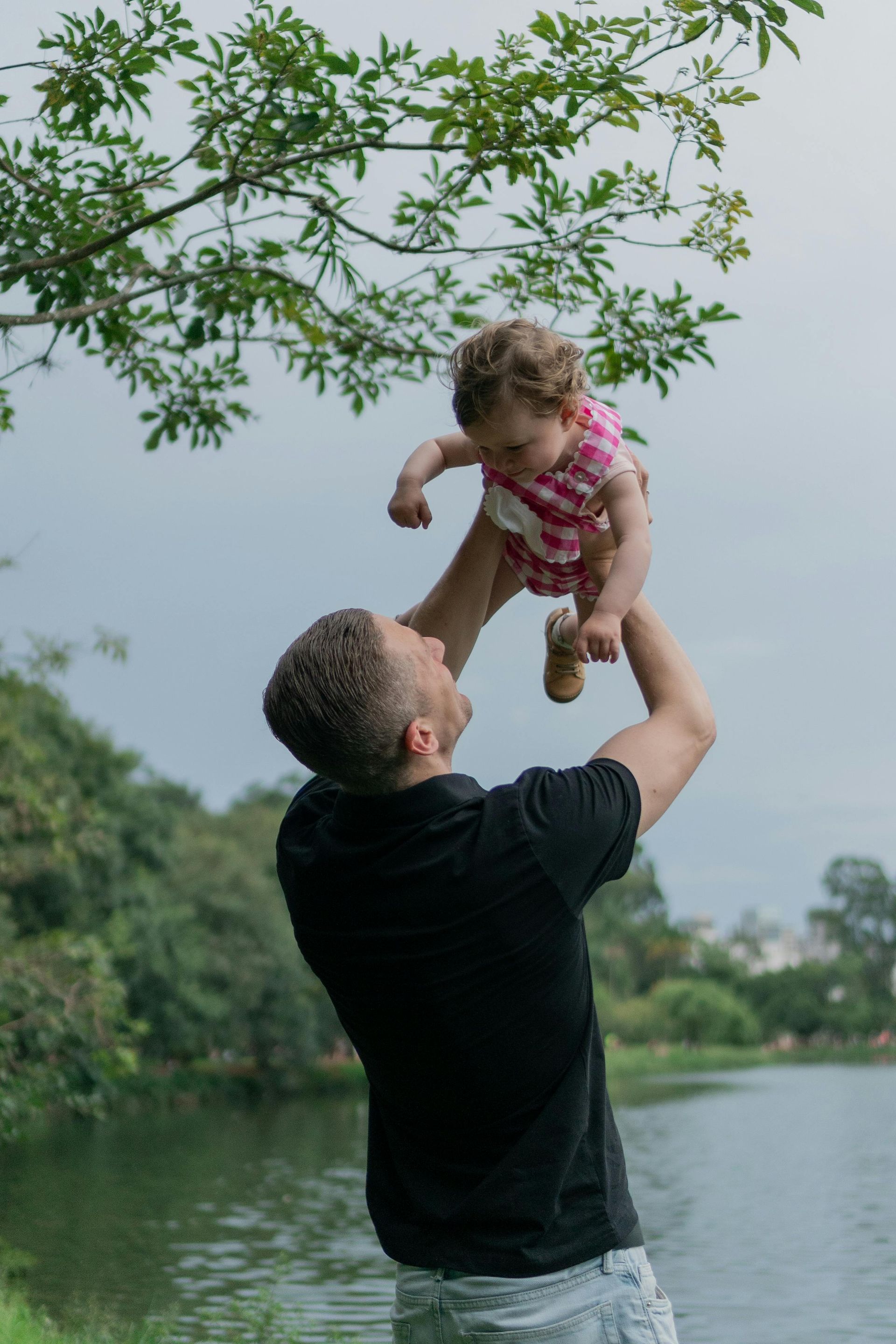 Man lifting a baby in the air near a lake with trees. Baby wears pink outfit.