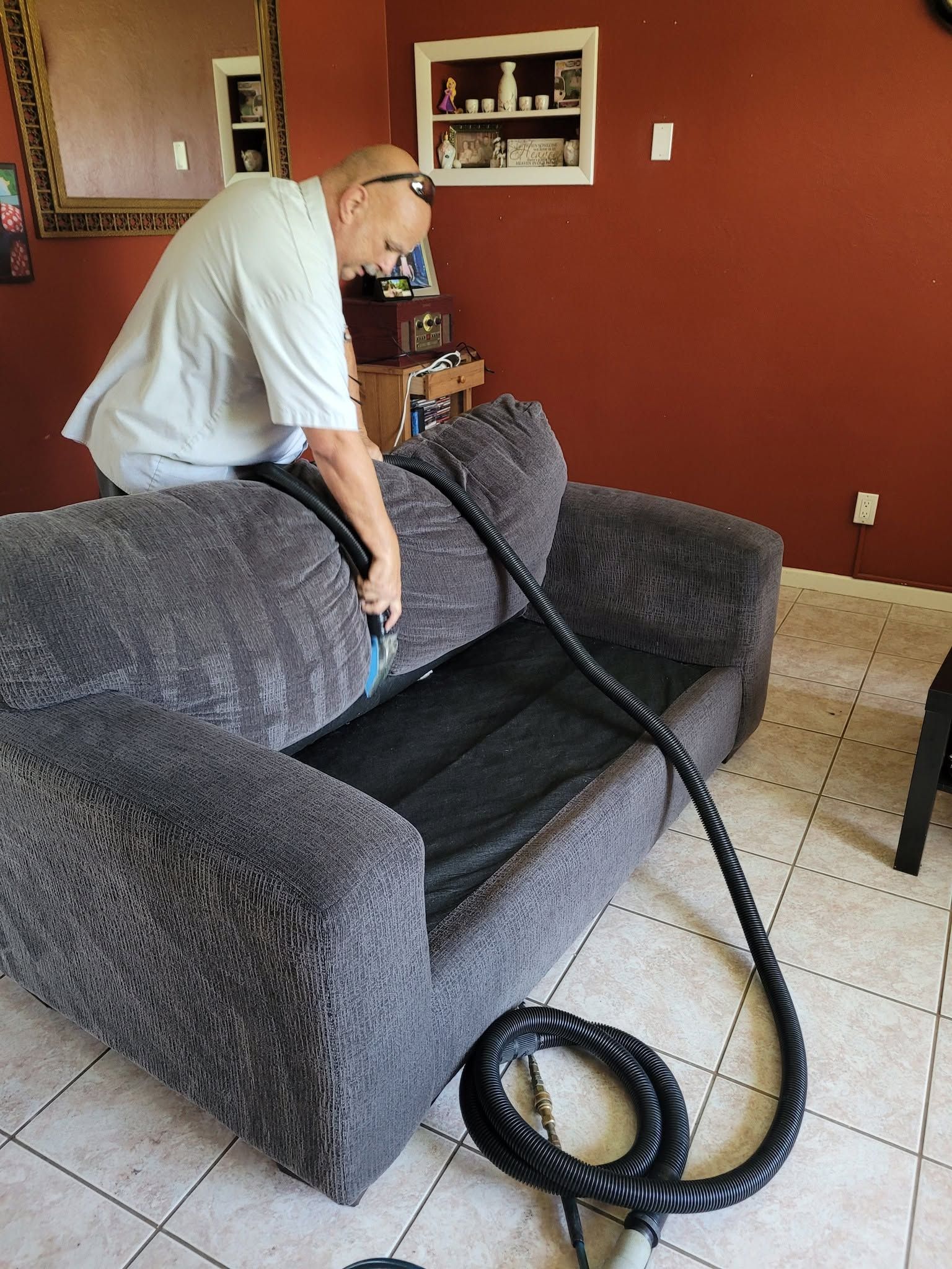 Man vacuuming a gray couch in a red-walled living room with a black hose on the floor