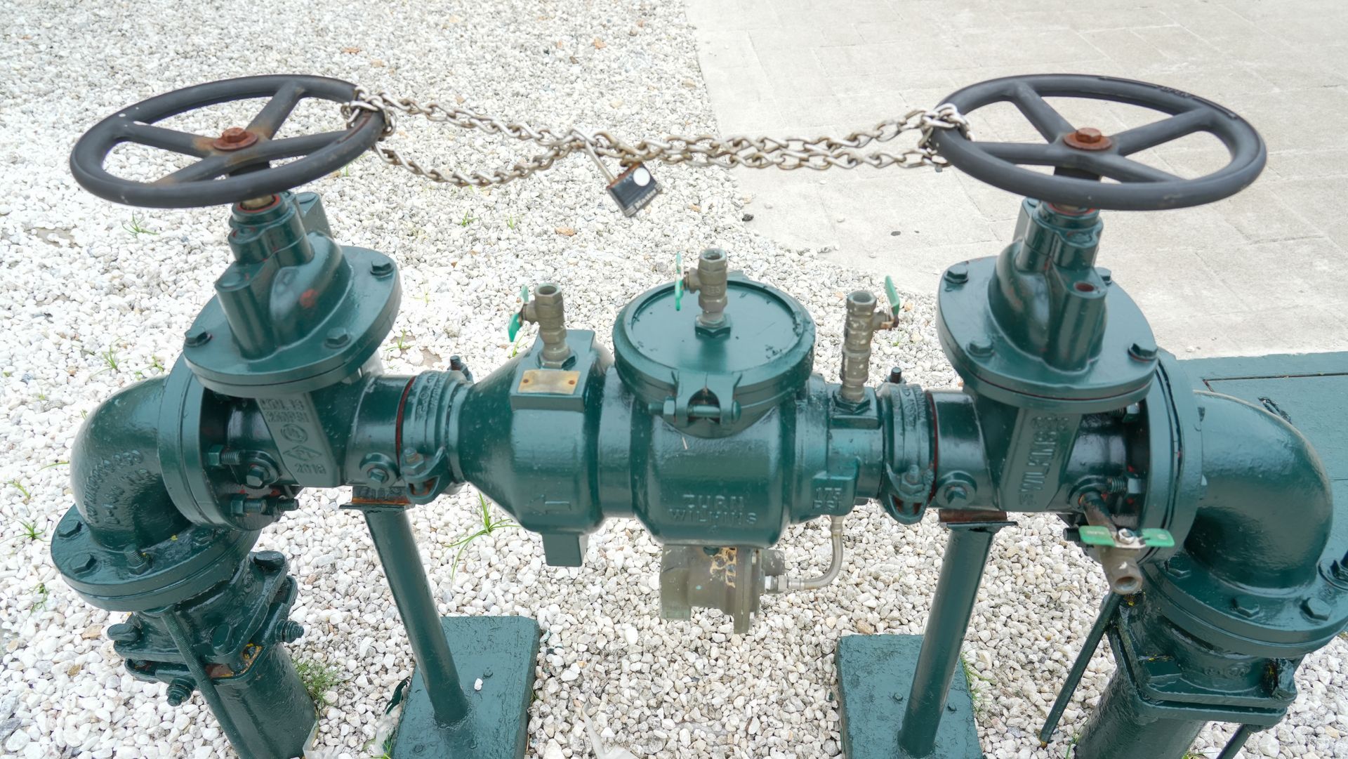 Green industrial pipeline with two black handwheel valves on a gravel surface