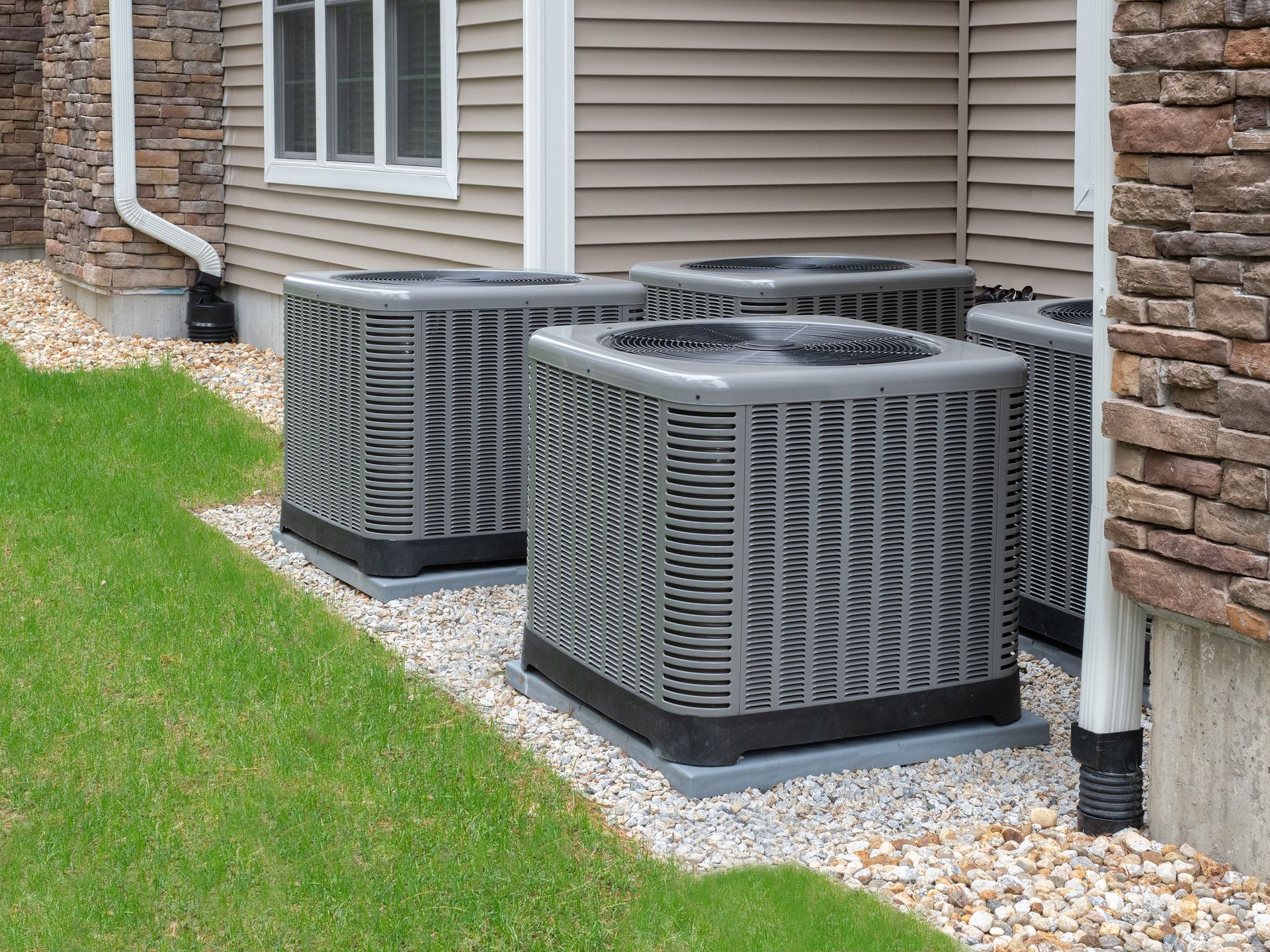 Two gray outdoor HVAC units beside a beige house wall and stone foundation on gravel and grass.