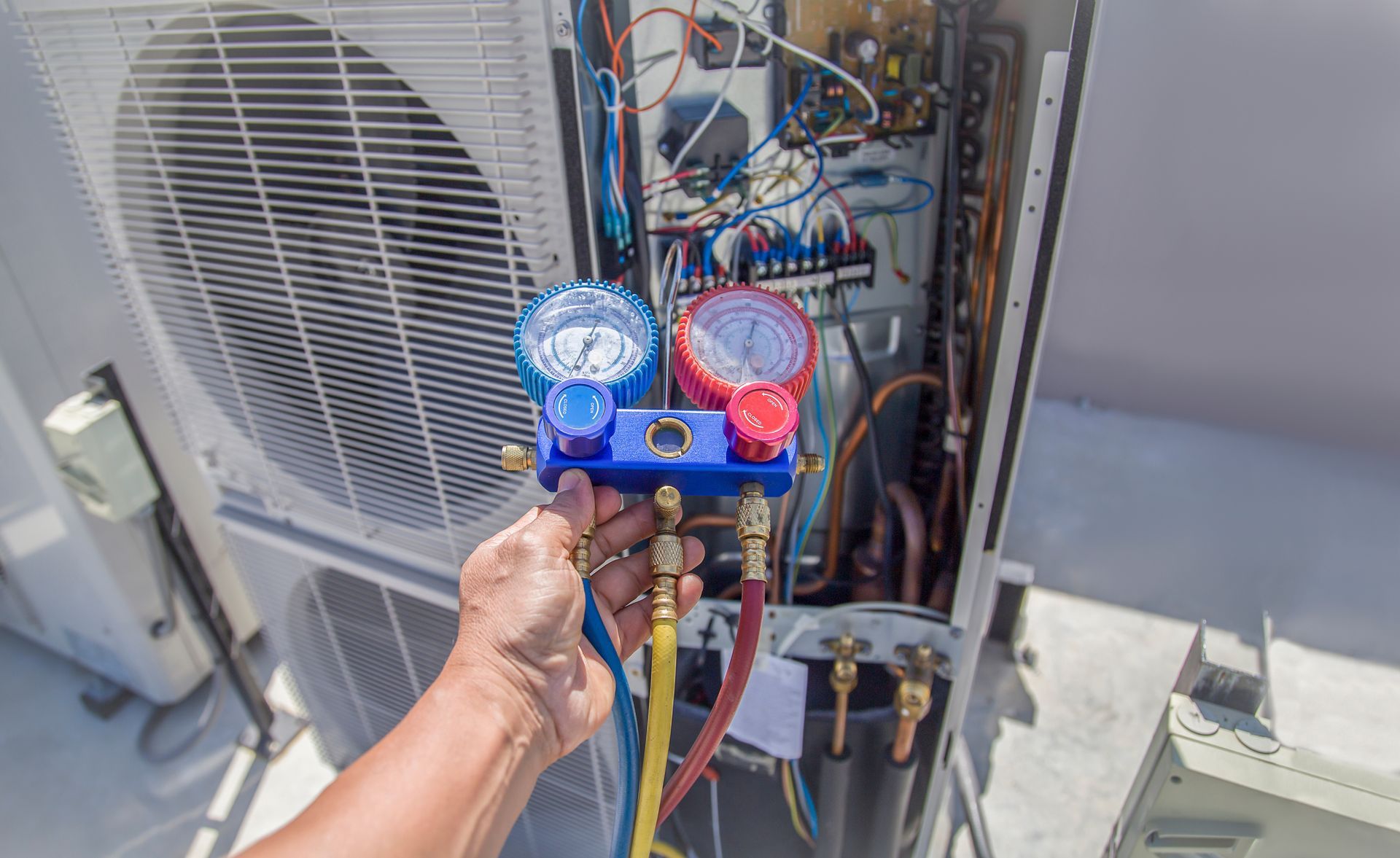 Hand holding HVAC manifold gauges in front of an open air-conditioning unit with exposed wiring and fans