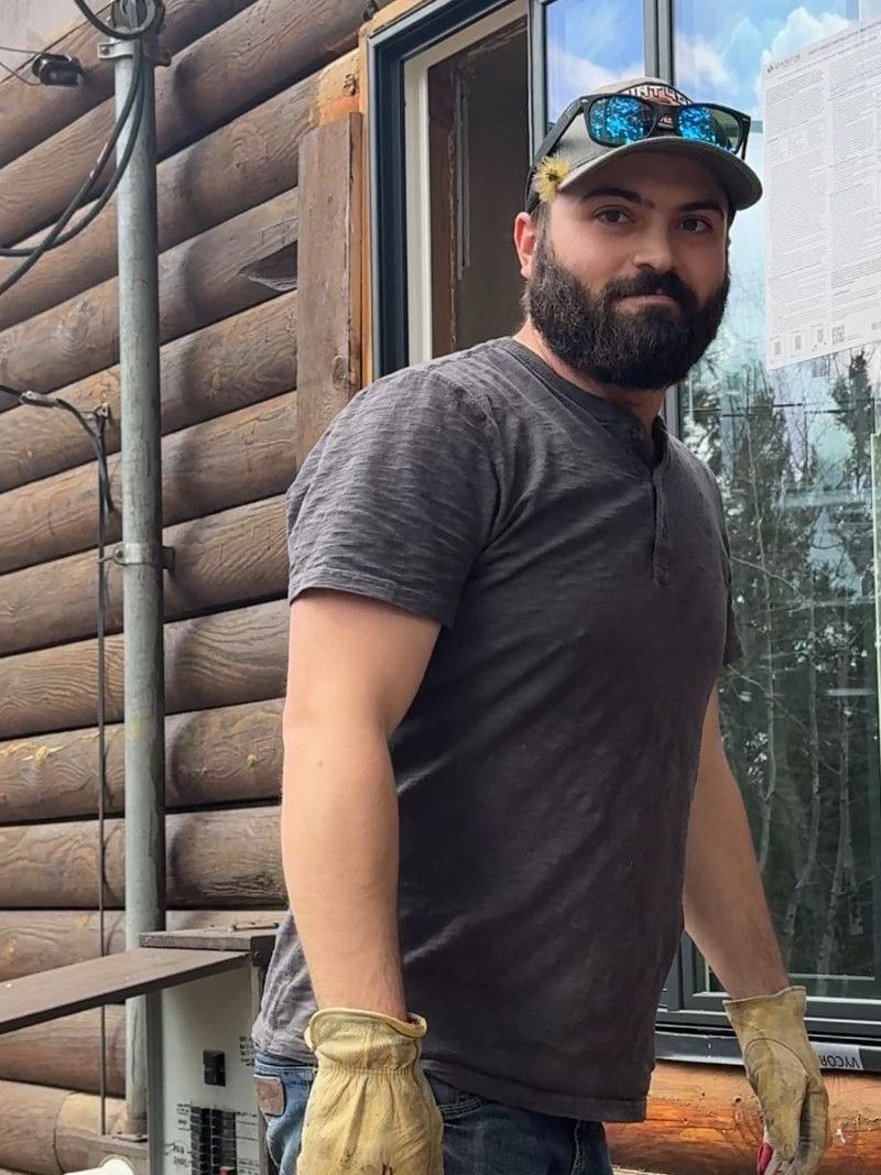 A man with a beard and hat is standing in front of a log cabin.