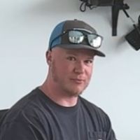 A man wearing a baseball cap and sunglasses is standing in front of a white wall.