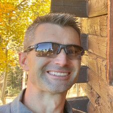 A man wearing sunglasses is smiling while leaning against a wooden wall.