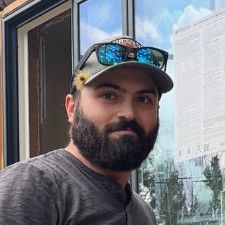 A man with a beard wearing a hat and sunglasses is standing in front of a window.