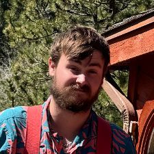 A man with a beard and suspenders is standing in front of a wooden building.