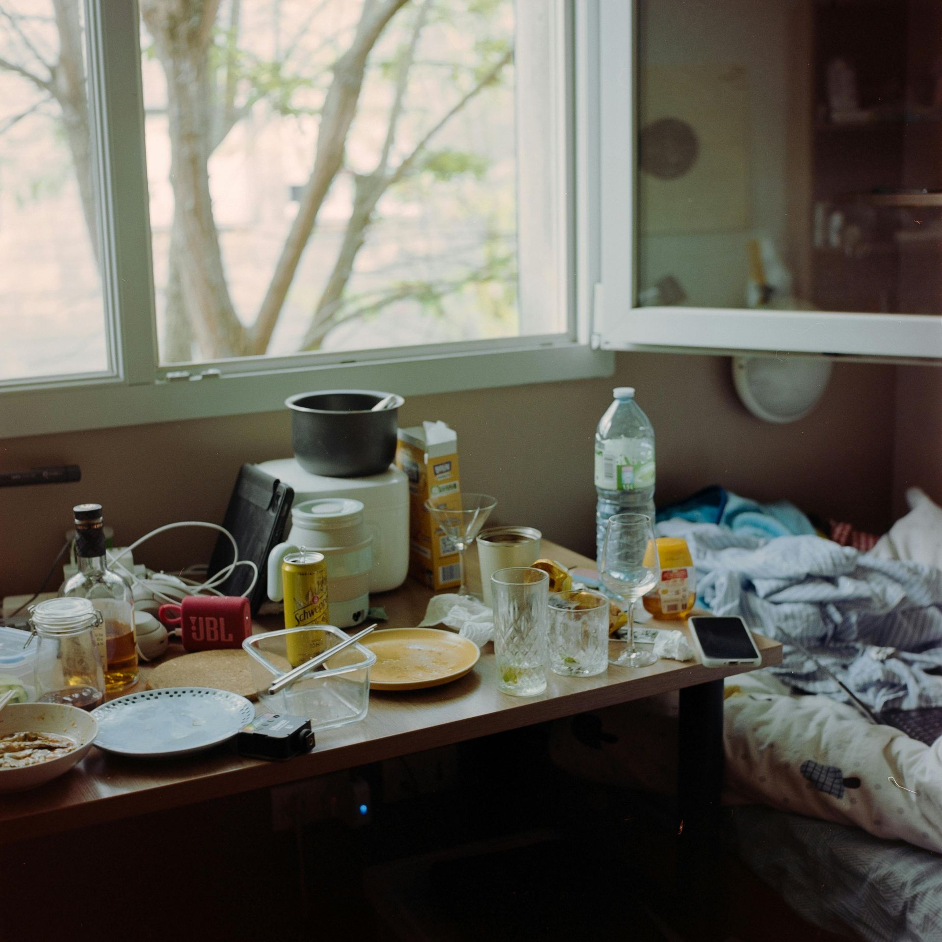 Messy tabletop near a window: food scraps, drinks, electronics, and water bottle. Sunlight streams in.