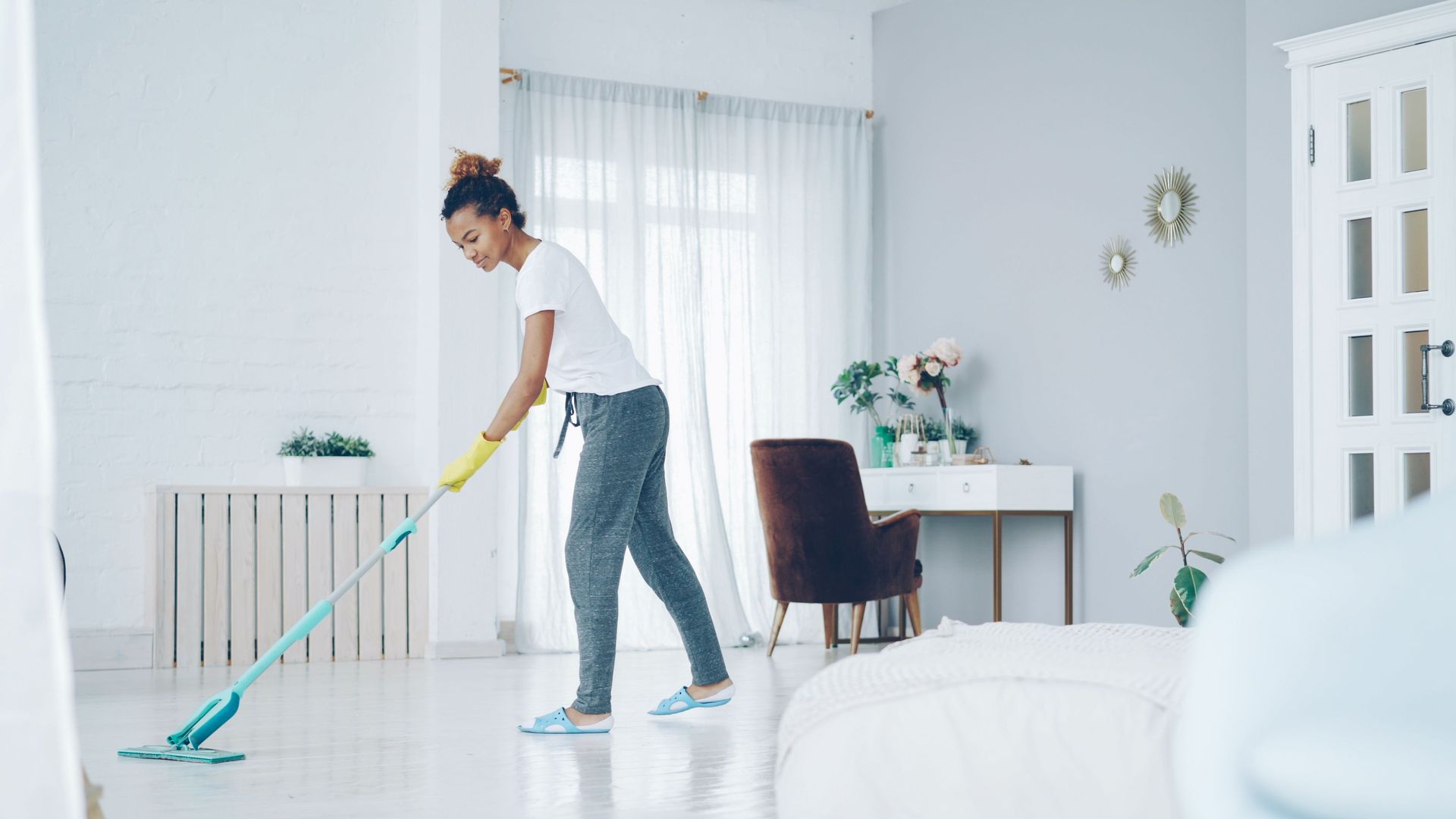 Woman mopping a bright white living room with turquoise mop and dark chair by a desk