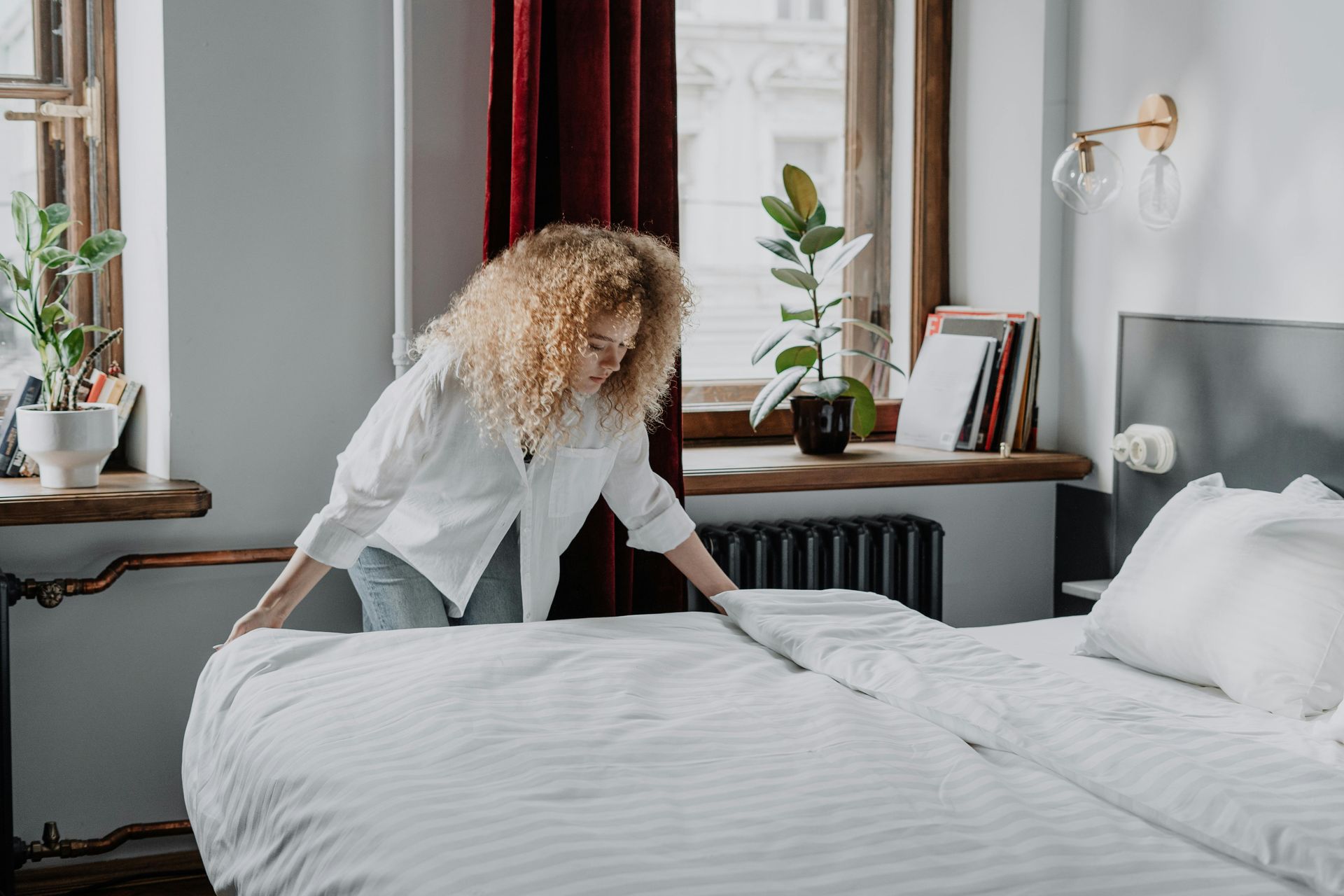 Woman making bed in a room with a window, plants, and red curtains.