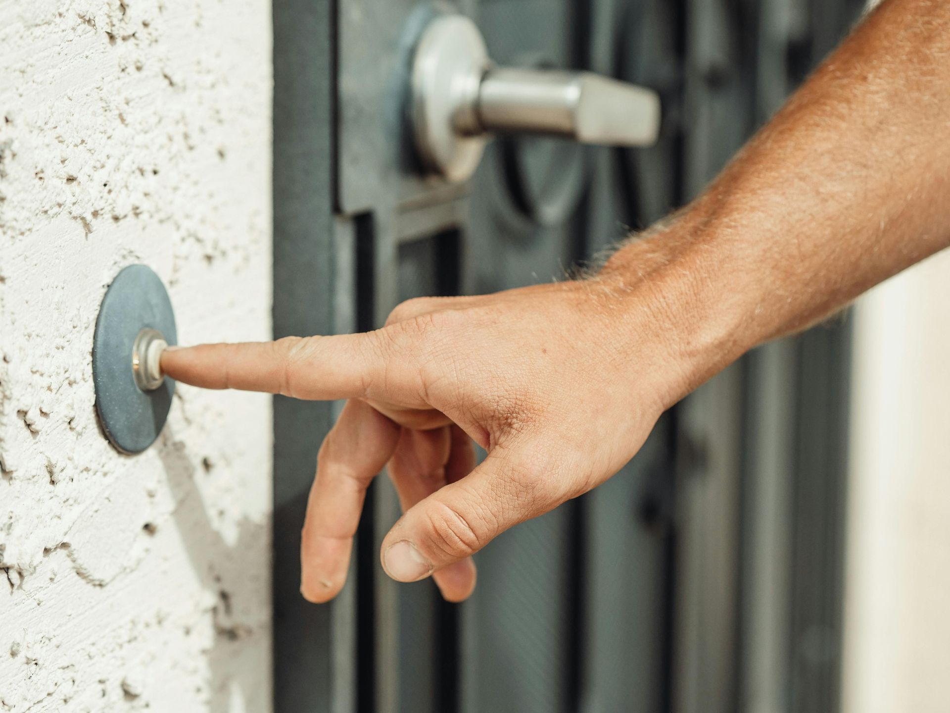 Person's finger pressing a doorbell button on a textured white wall near a closed gray door.