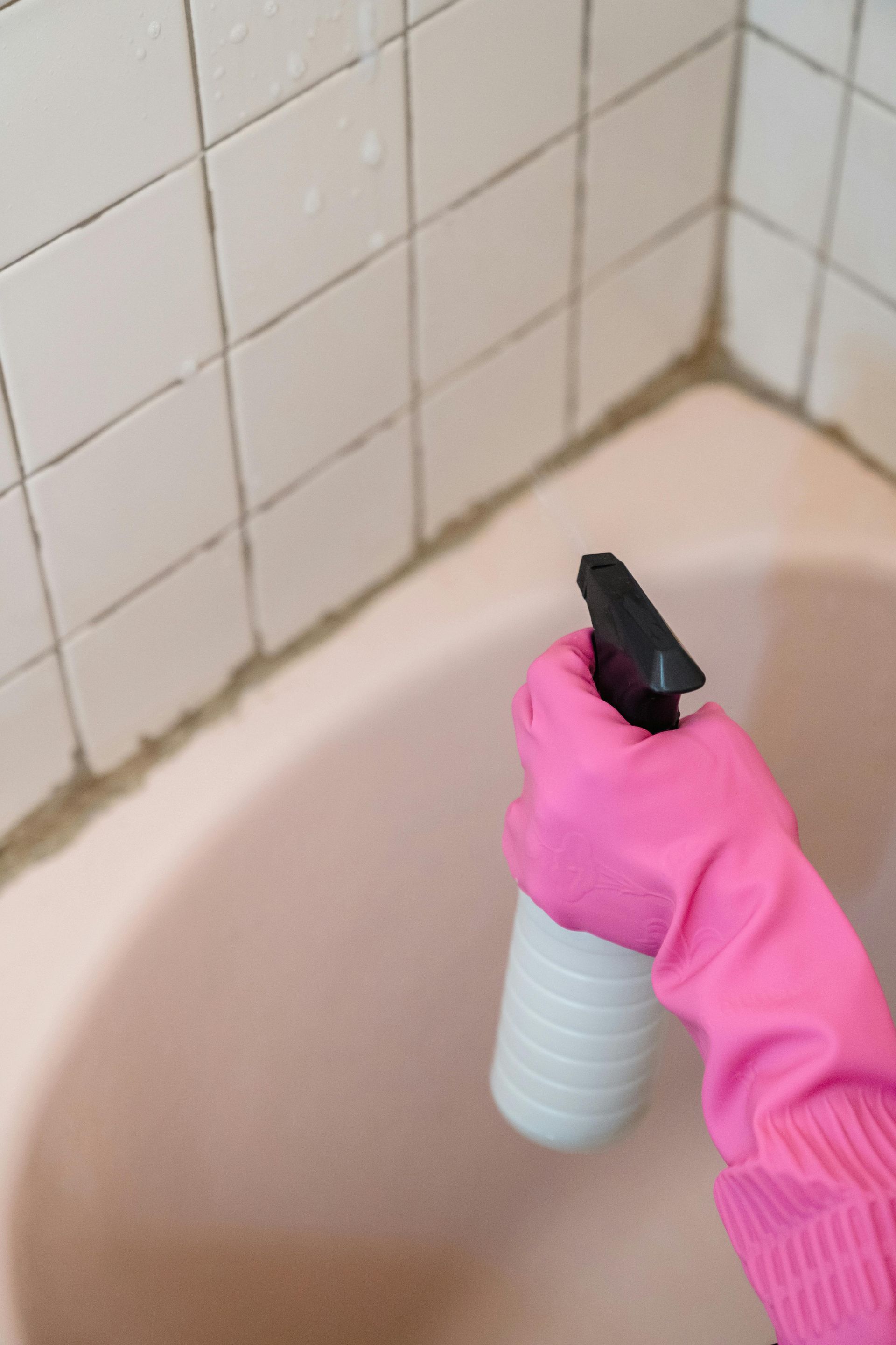 Hand in pink glove sprays a white bottle at a pink bathtub with tiled walls.