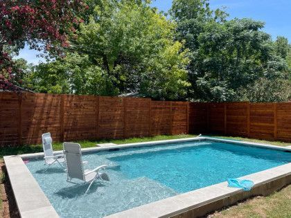 A rectangular swimming pool with two chairs, surrounded by a wooden fence and greenery.