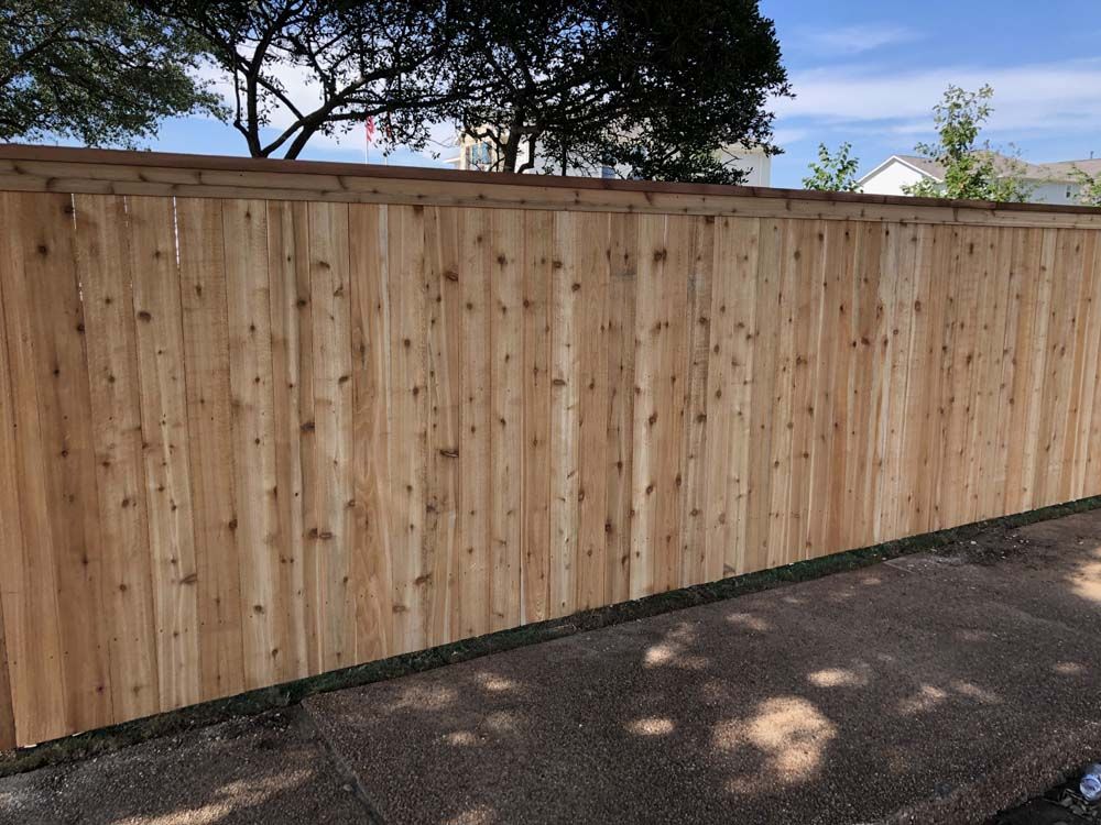 Wooden fence along a paved surface with trees in the background.
