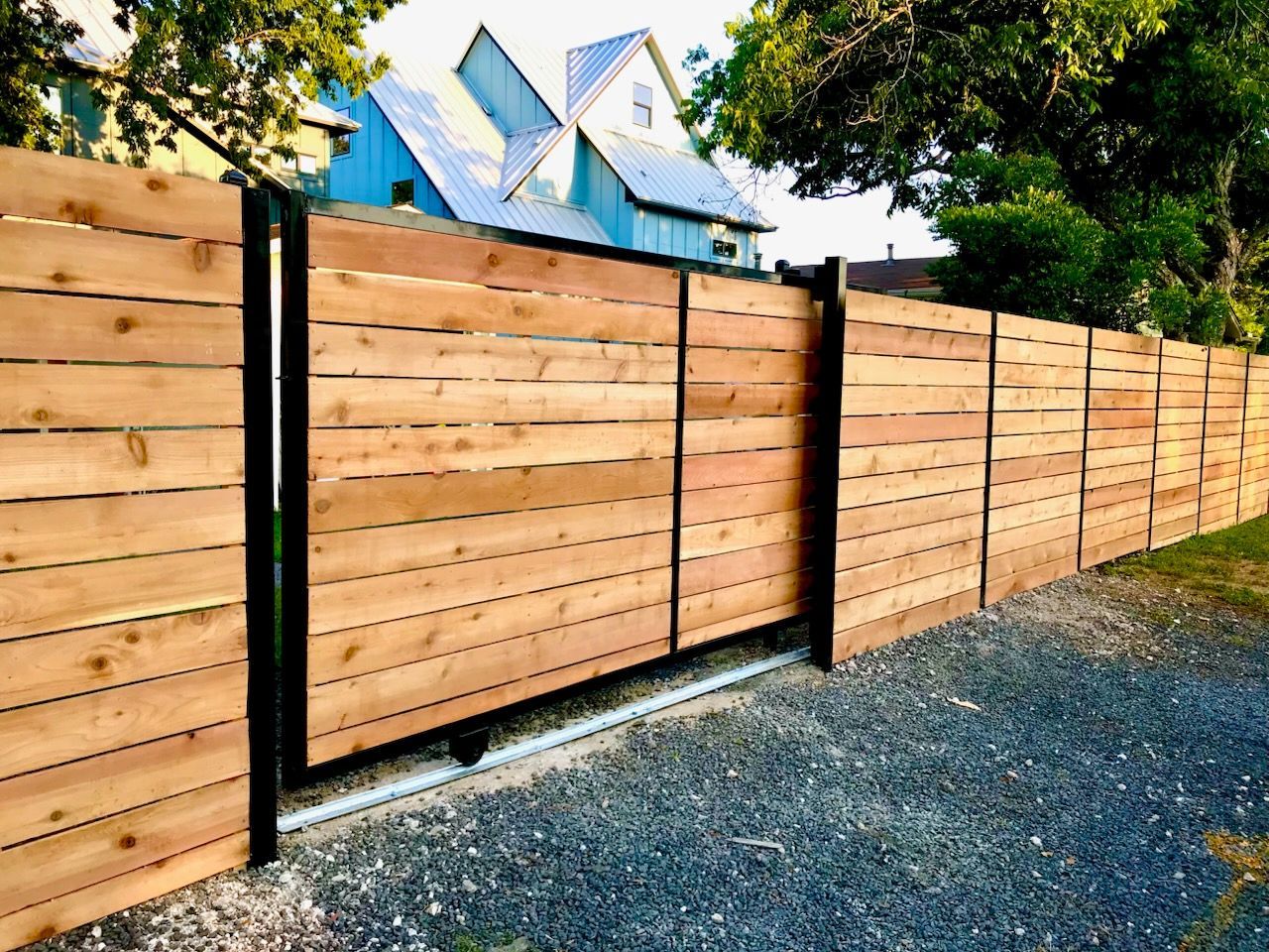 Wooden horizontal slat fence with black metal posts and gate, gravel driveway, house in background.