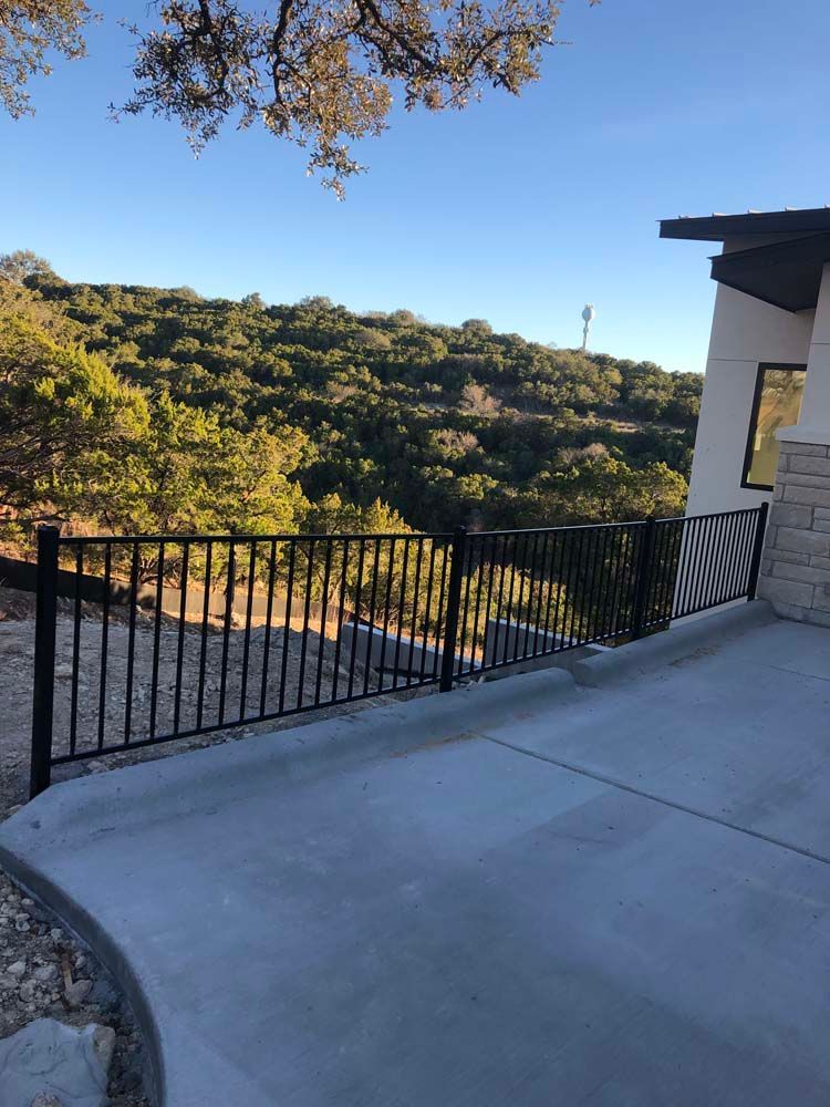 Black metal fence bordering a concrete patio, with a hillside and a building in the background.