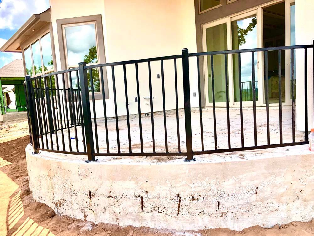 Black metal railing on a concrete patio overlooking a sandy area, near a house with large windows.