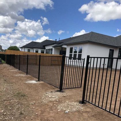 Black metal fence enclosing a backyard with a house, blue sky, and clouds.