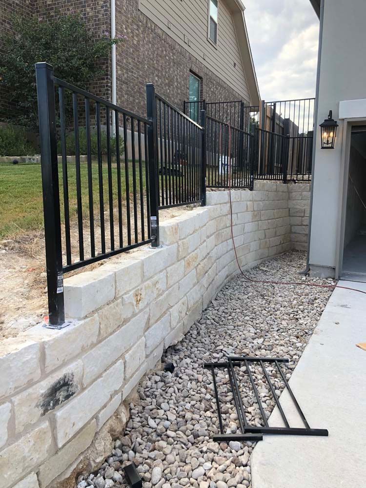 Black metal fence on a light stone retaining wall next to a house with gravel.