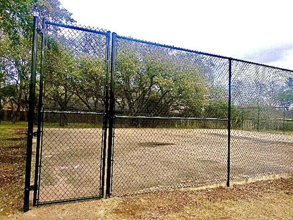 Black chain-link fence with a double-door gate, enclosing an empty dirt area, in a grassy park.