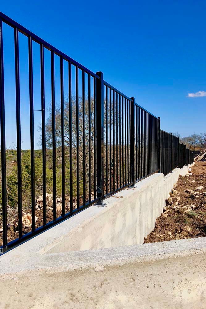 Black metal fence atop a concrete barrier, under a blue sky, overlooking a natural landscape.