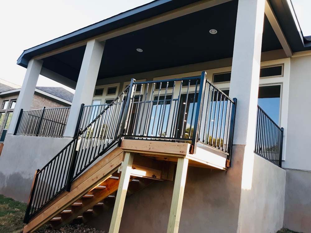 Black railing surrounds a wooden deck with stairs, attached to a house with beige walls and a dark roof.