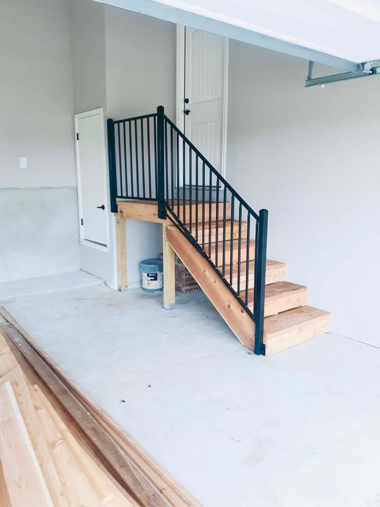 Wooden staircase with black railing in a garage. Grey walls and a white door are visible.