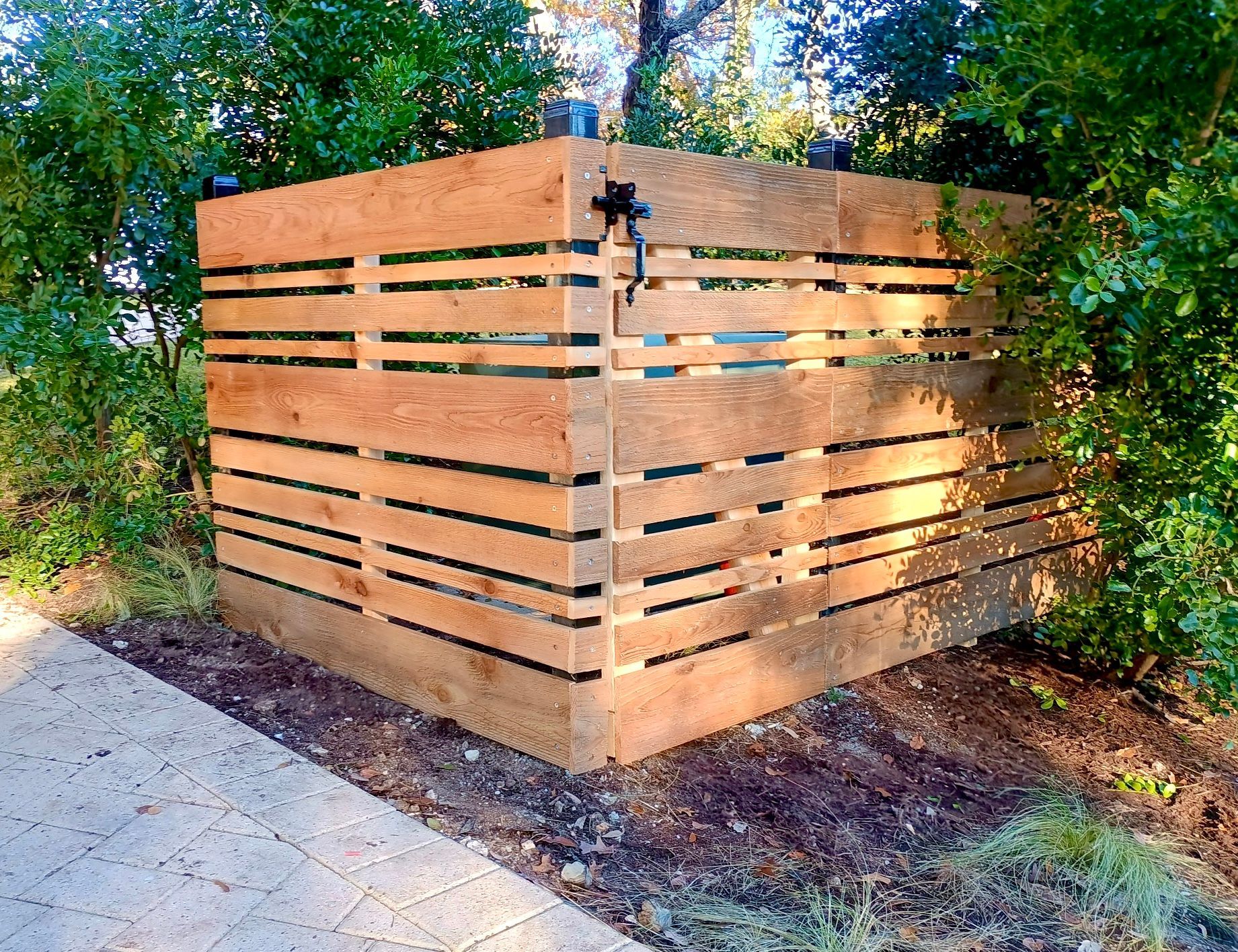 Wooden enclosure with horizontal slats, concealing equipment in a garden setting, partially obscured by bushes.