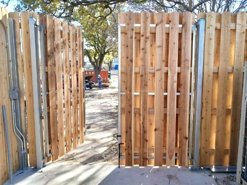 Open wooden gate in a wooden fence; beyond, a construction area and people.