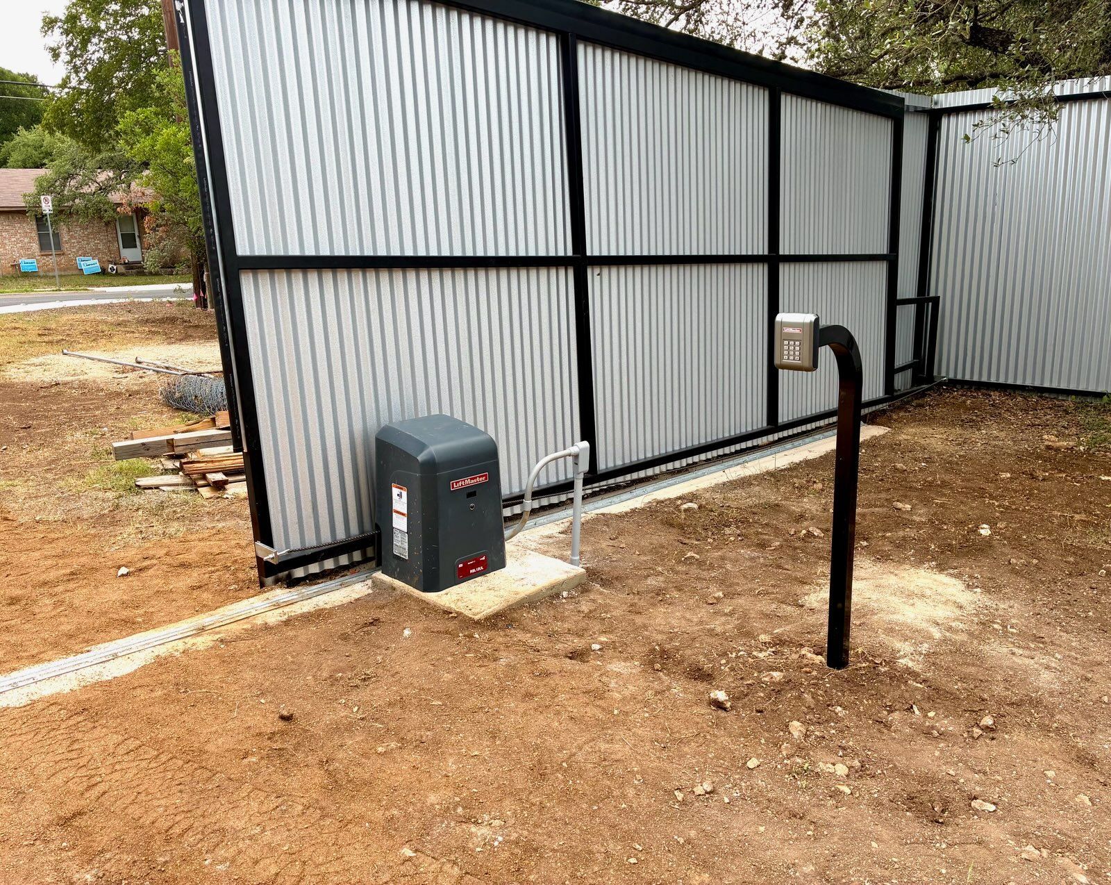 Metal gate with automatic opener and access controls, set in a yard with gravel and a nearby building.