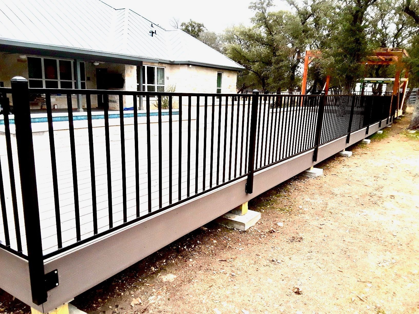 Black metal railing surrounds a gray deck next to a pool and a light-colored house.