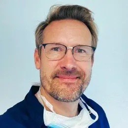 Tannlege Christian med briller og skjegg, iført blå skjorte og medisinsk maske, smiler til kameraet.