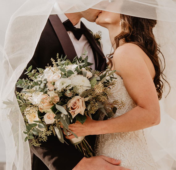 Una novia y un novio besándose bajo un velo mientras la novia sostiene un ramo de flores