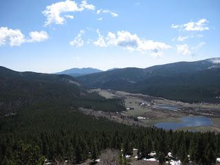 A high-angle view of a mountain valley with a pine forest in the foreground, small lakes, and distant blue mountain peaks.