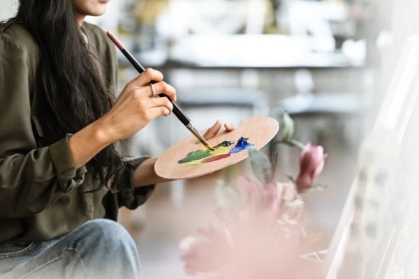A person in an olive shirt holds a wooden palette and paintbrush, preparing to paint on a canvas.