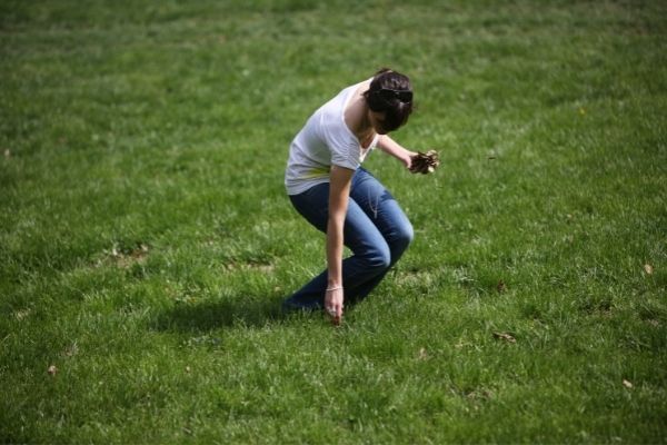 A person in a white shirt and blue jeans bends over to pick something up from a grassy lawn.