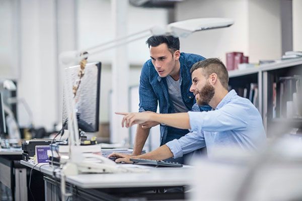 Two colleagues at a desk look at a computer screen, one pointing at the monitor as they collaborate in an office.