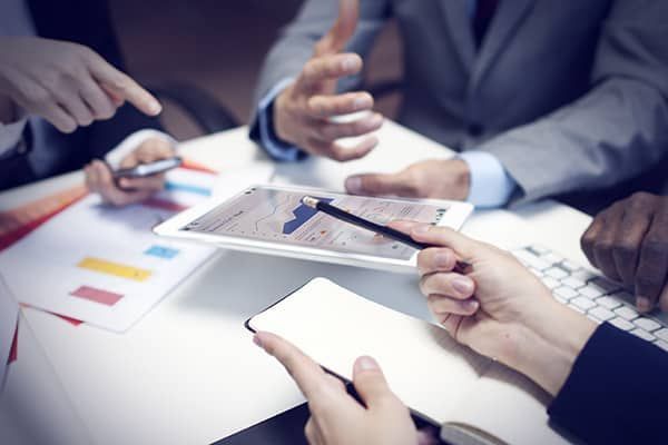 Business meeting at a white table, people pointing at charts on a tablet and using phones.