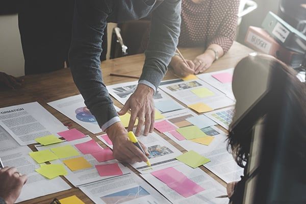 People gathered around a table with papers, sticky notes, and a pen, discussing a project.Systems Optimization: Your process might not look the same with you doing it versus you giving it to somebody else to do it.