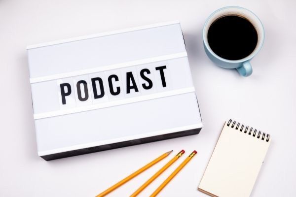 Lightbox displaying the word PODCAST, with a coffee mug, notepad, and pencils on a white surface.