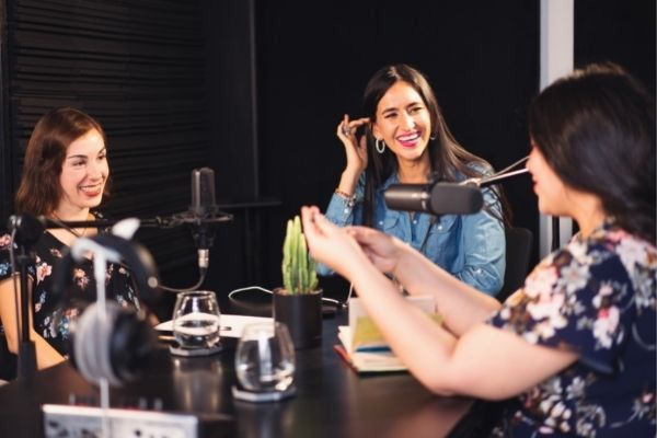 Three women laughing while recording a podcast in a studio with microphones and equipment.