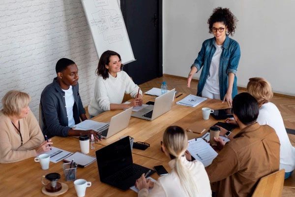 A group of people in an office meeting around a wooden table. A woman is presenting while others take notes.