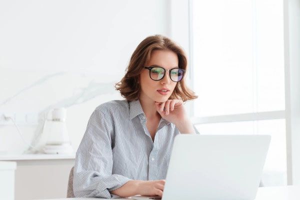 Woman with glasses, resting chin on hand, looking at laptop in light-filled room.