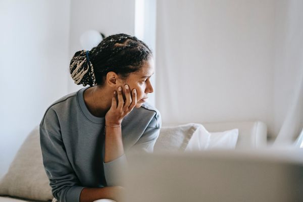 Woman with hand on cheek, looking off to the side, seated on a couch in a bright room.