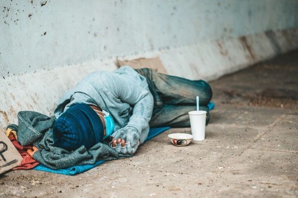 Person sleeping on the ground under a bridge with a cup and bowl next to them